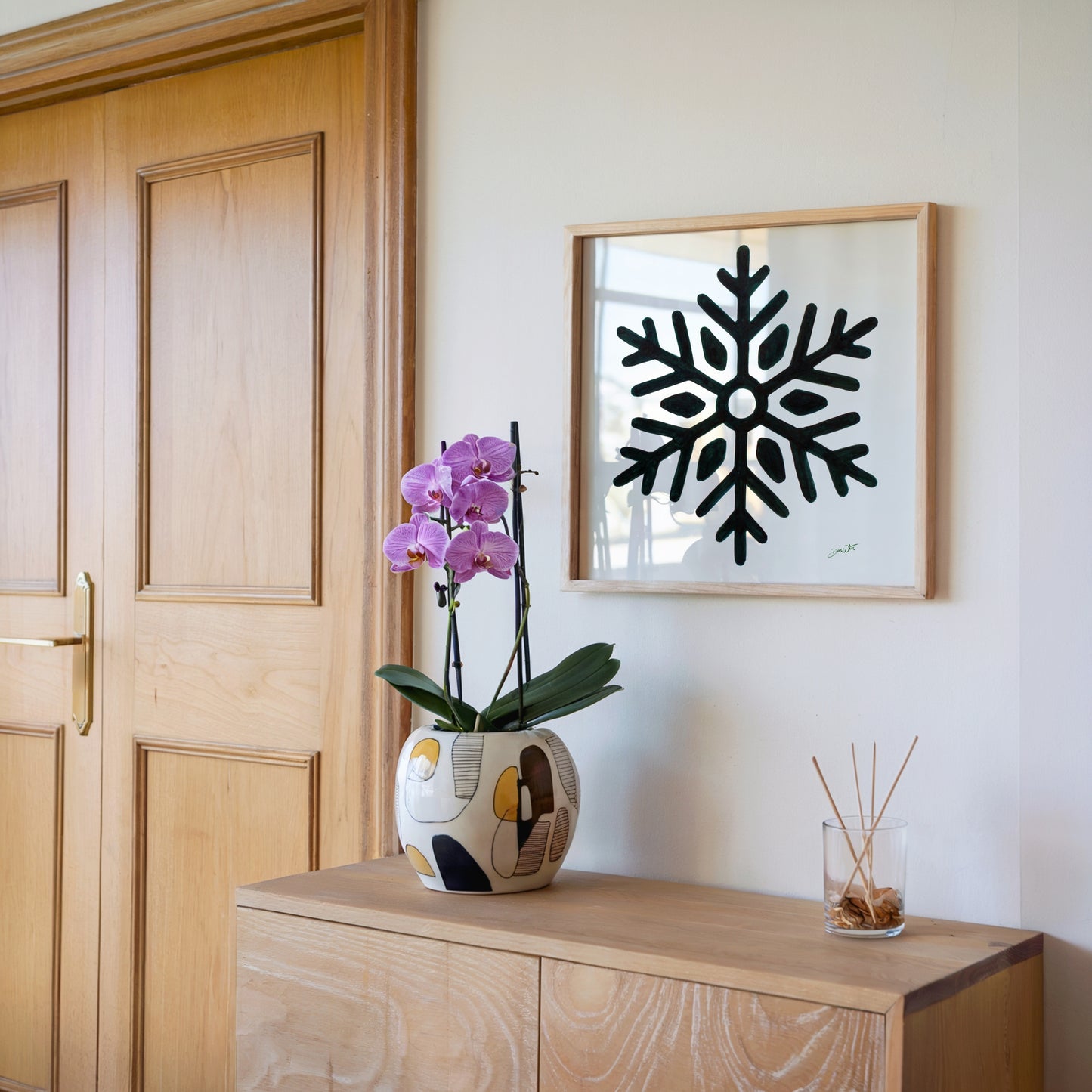 Decorative setup with a vase of flowers, a diffuser, and a framed green snowflake art piece on a wooden surface.