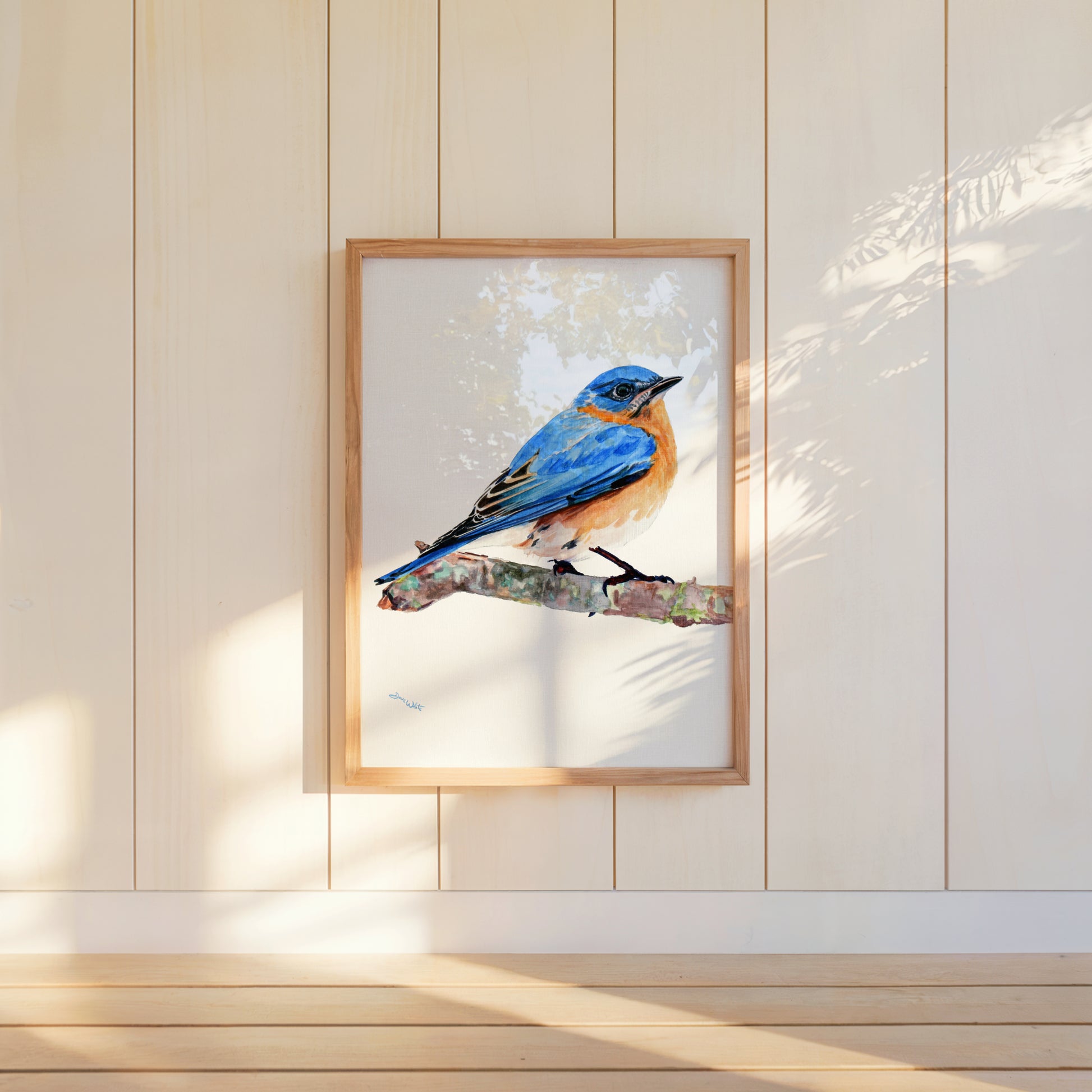Framed artwork of a blue bird on a branch against a light wooden panel wall.