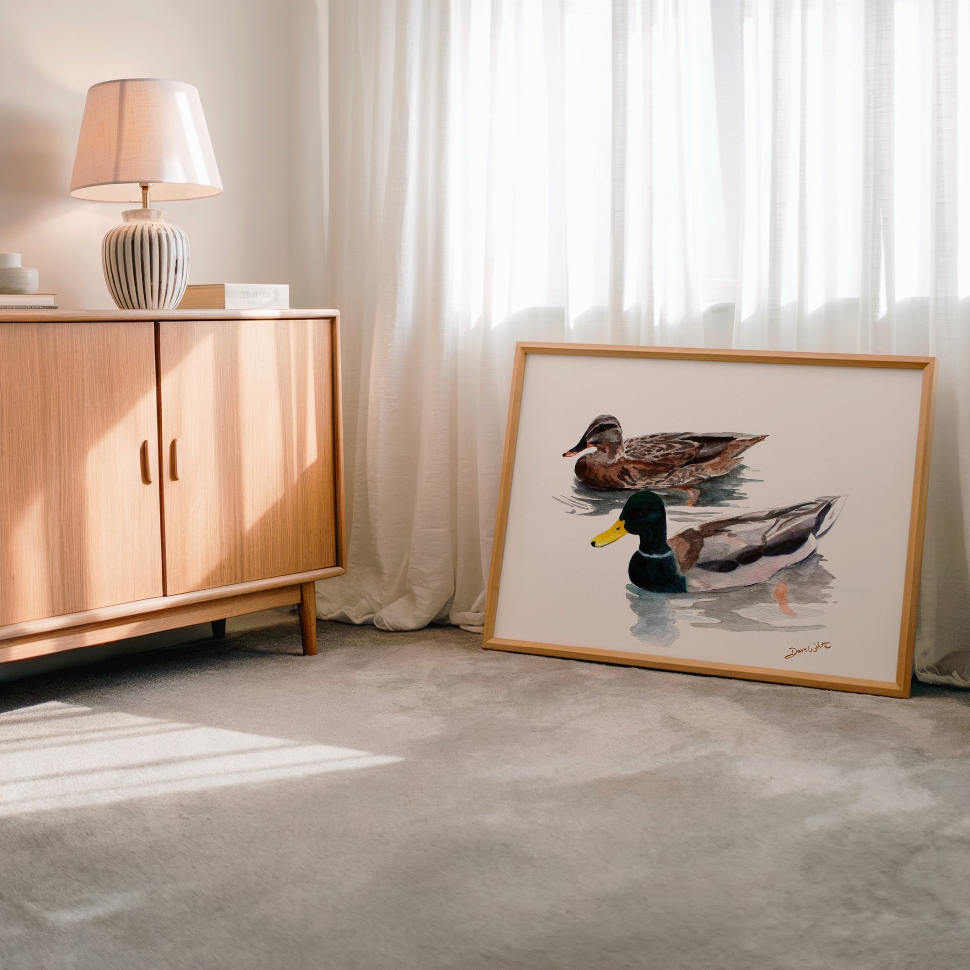 Framed artwork of ducks on a floor next to a wooden cabinet with a lamp.