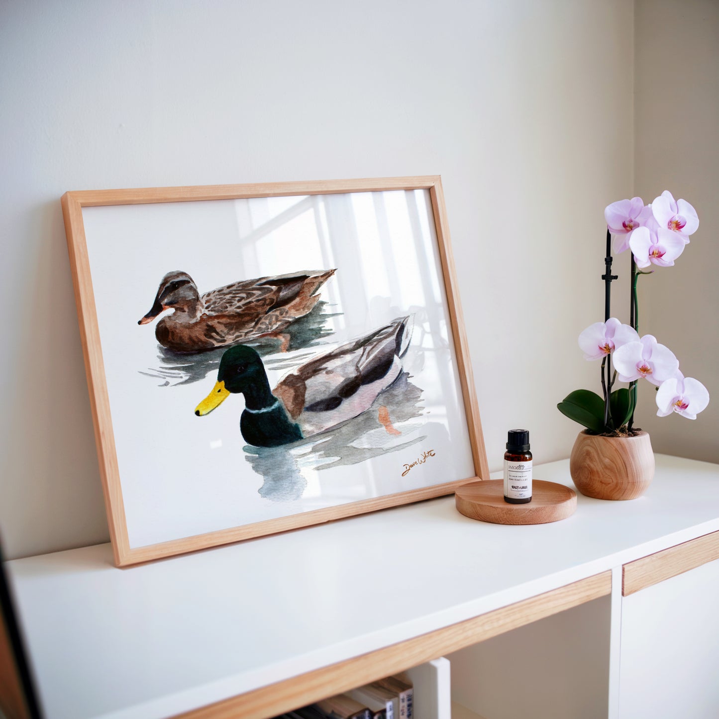 Framed artwork of ducks on a shelf with a potted orchid and small bottle.